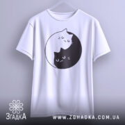Футболка з малюнком котів білого кольору, підвішена на вішалці, з принтом двох котів, один чорний, а інший білий, що обіймаються в кругу. Фон світло-сірий, що надає контрасту принту.