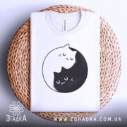 Футболка з малюнком котів білого кольору, складена на кошику з ротанга, з принтом двох котів, один чорний, а інший білий, що обіймаються в кругу. Фон світло-сірий, що підкреслює текстуру кошика.
