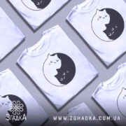 Футболка з малюнком котів білого кольору, складена в ряд, з принтом двох котів, один чорний, а інший білий, що обіймаються в кругу. Фон світло-сірий, з кількома футболками, що створює враження колекції.