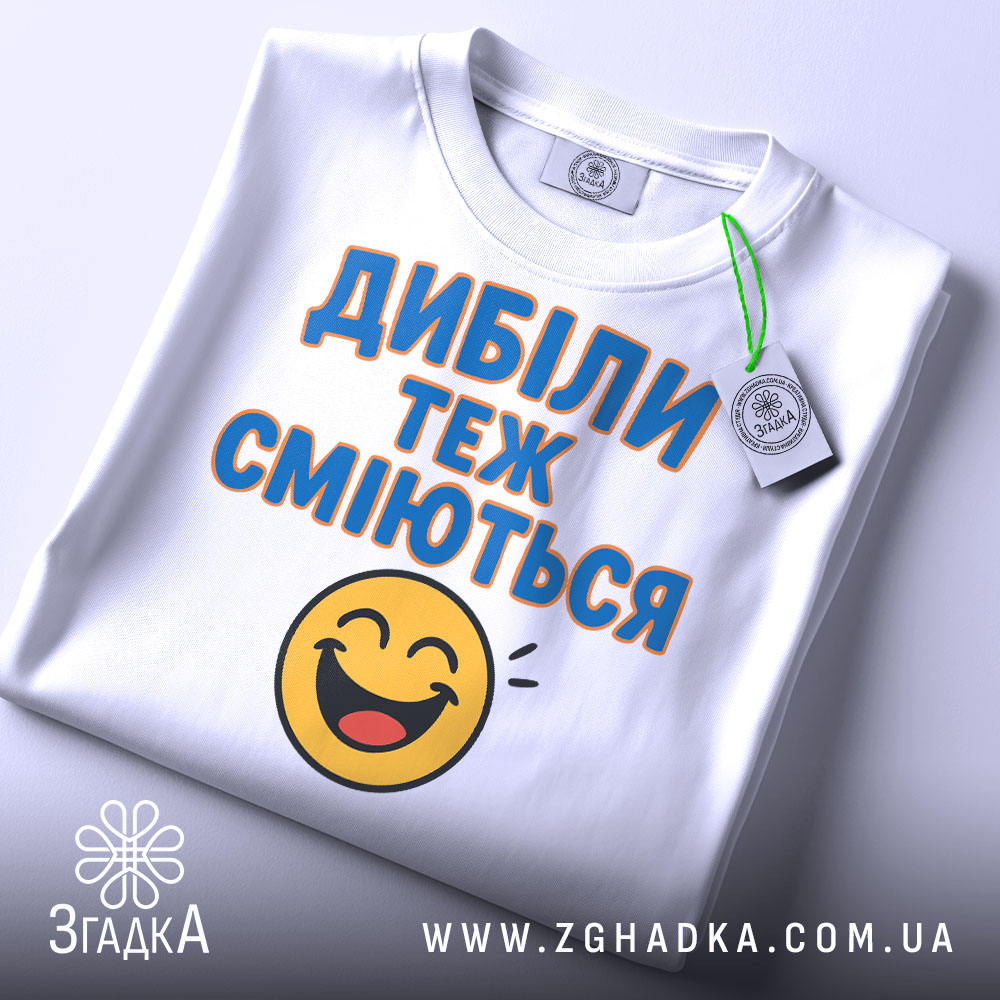 Футболка з написом Дибіли, білого кольору, з верхнього ракурсу, складена на поверхні, фон світлий, принт з текстом "Дибіли теж сміються" та смайлом, з етикеткою.