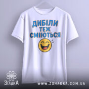 Футболка з написом Дибіли, білого кольору, з фронтального ракурсу, підвішена на вішалці, фон нейтральний, принт з текстом "Дибіли теж сміються" та смайлом.