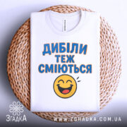 Футболка з написом Дибіли, білого кольору, з верхнього ракурсу, складена на кошику, фон світлий, принт з текстом "Дибіли теж сміються" та смайлом.