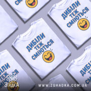 Футболка з написом Дибіли, білого кольору, з верхнього ракурсу, кілька футболок розкладені на поверхні, фон світлий, принт з текстом "Дибіли теж сміються" та смайлом.