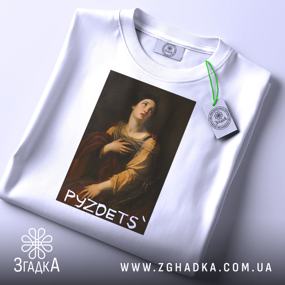 Футболка з написом PYZDETS, білого кольору, лежить на поверхні, фон світлий, принт зображає жінку в історичному костюмі, що дивиться вгору, з червоним плащем, на комірці є етикетка.