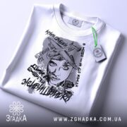 Футболка з написом свободи, білого кольору, складена на плоскій поверхні, фон світло-сірий, принт зображує жінку з літаками та текстами, на футболці є зелений ярлик.