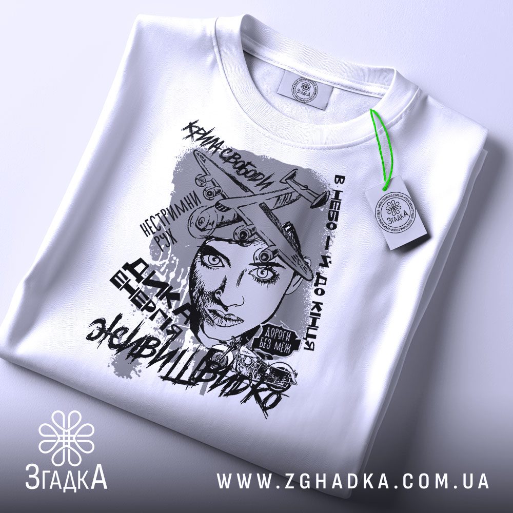 Футболка з написом свободи, білого кольору, складена на плоскій поверхні, фон світло-сірий, принт зображує жінку з літаками та текстами, на футболці є зелений ярлик.