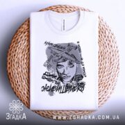 Футболка з написом свободи, білого кольору, складена на плоскій поверхні з плетеного матеріалу, фон світло-сірий, принт містить зображення жінки з літаками та текстами.