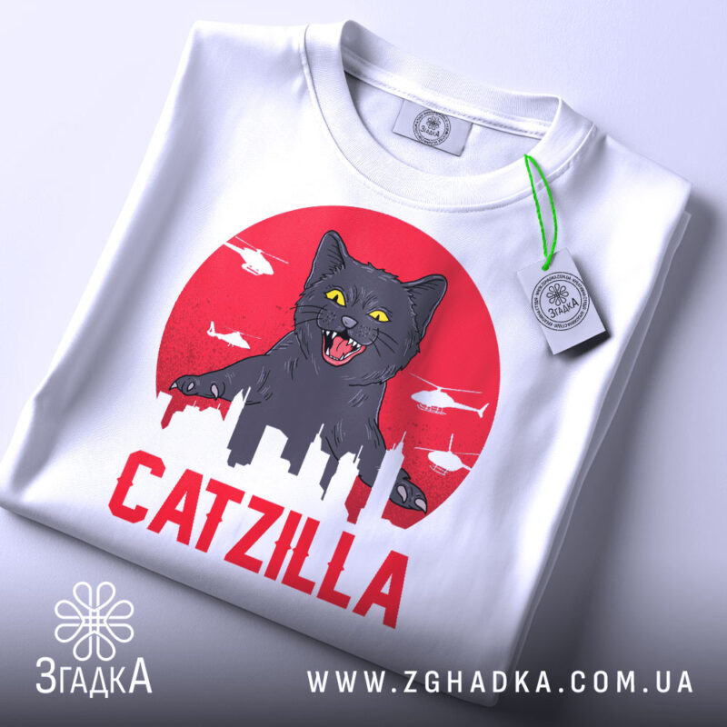 Футболка з принтом Catzilla білого кольору, зображена зверху, на ній є етикетка, фон світло-сірий, принт показує чорного кота на червоному фоні з силуетами міста та вертольотами.
