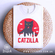 Футболка з принтом Catzilla білого кольору, зображена зверху на плетеній основі, фон світло-сірий, принт показує чорного кота на червоному фоні з силуетами міста та вертольотами.