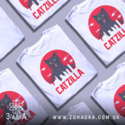 Футболка з принтом Catzilla білого кольору, зображена з фронтального ракурсу, на ній кілька футболок, фон світло-сірий, принт містить чорного кота на червоному фоні з силуетами міста та вертольотами.
