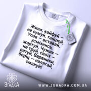Футболка живи кайфуй не сумуй. Біла футболка, складена на поверхні, з етикеткою, що звисає. Текст чорного кольору чітко видимий, фон нейтральний.