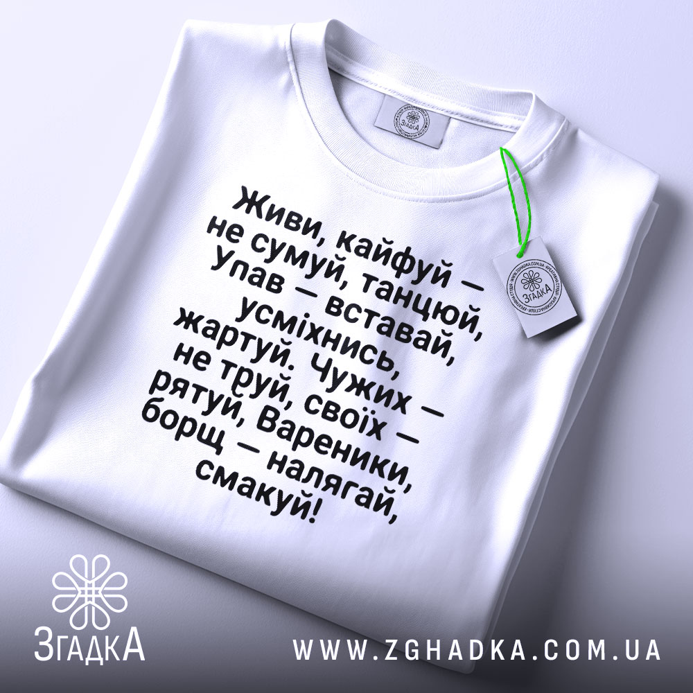 Футболка живи кайфуй не сумуй. Біла футболка, складена на поверхні, з етикеткою, що звисає. Текст чорного кольору чітко видимий, фон нейтральний.