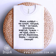 Футболка живи кайфуй не сумуй. Біла футболка, складена на круглій кошику, з текстом чорного кольору. Фон натуральний, з акцентом на текст.