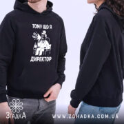 Худі я директор подарунок. Чорний колір, ракурс з боку, фон світлий. Дві людини в худі, один з них тримає кишеню-«кенгуру», принт з малюнком директора з текстом "ТОМУ ЩО Я ДИРЕКТОР". Капюшони видимі, манжети на руках.