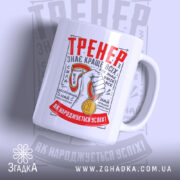 Чашка для найкращого тренера, біла, зображена в ракурсі з правого боку, на фоні світло-сірого кольору. Принт містить червоний напис 'ТРЕНЕР' та зображення руки з медаллю. Ручка кругла, край чашки гладкий, поверхня матова.