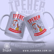 Чашка для найкращого тренера, біла, зображена в ракурсі з двох боків, на фоні світло-сірого кольору. Принт містить червоний напис 'ТРЕНЕР' та зображення руки з медаллю. Ручки чашок круглі, краї гладкі, поверхня матова.