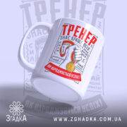 Чашка для найкращого тренера, біла, зображена в ракурсі з правого боку, на фоні світло-сірого кольору. Принт містить червоний напис 'ТРЕНЕР' та зображення руки з медаллю. Ручка кругла, край чашки гладкий, поверхня матова.