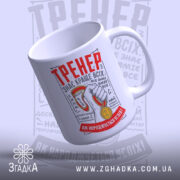 Чашка для найкращого тренера, біла, зображена в ракурсі з лівого боку, на фоні світло-сірого кольору. Принт містить червоний напис 'ТРЕНЕР' та зображення руки з медаллю. Ручка кругла, край чашки гладкий, поверхня матова.