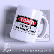 Чашка геній за роботою. Чашка білого кольору з чорним і червоним принтом, на якому написано 'УВАГА ГЕНІЙ ЗА РОБОТОЮ'. Ручка гладка, край рівний, поверхня має легкий глянець. Фон світло-фіолетовий, з розмитим логотипом.