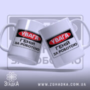 Чашка геній за роботою. Дві чашки білого кольору з однаковим принтом 'УВАГА ГЕНІЙ ЗА РОБОТОЮ', розташовані під кутом одна до одної. Ручки гладкі, краї рівні, поверхня має легкий глянець. Фон світло-фіолетовий, з розмитим логотипом.