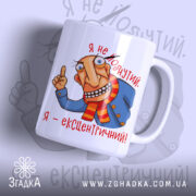 Чашка я ексцентричний, біла з кольоровим малюнком, показує персонажа з великими очима та усмішкою, що одягнений у синю куртку і червону шарф, ручка з правого боку, край гладкий, глянцевий фініш.