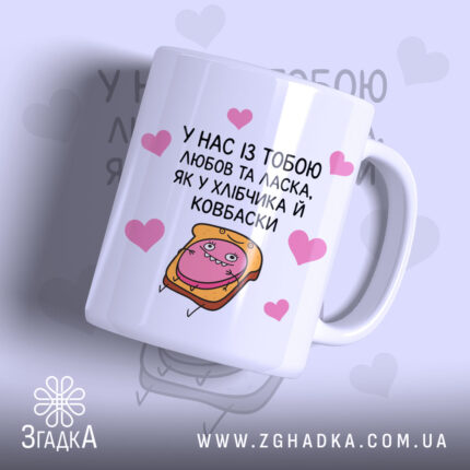Чашка хлібчик й ковбаска, біла з кольоровим принтом, показана з правого ракурсу, на фоні світло-фіолетового кольору з сердечками, з малюнком хлібчика і ковбаски, ручка класична, край рівний, глянцева поверхня.