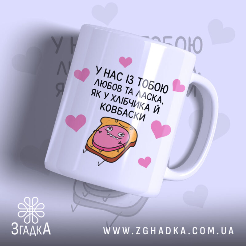 Чашка хлібчик й ковбаска, біла з кольоровим принтом, показана з правого ракурсу, на фоні світло-фіолетового кольору з сердечками, з малюнком хлібчика і ковбаски, ручка класична, край рівний, глянцева поверхня.
