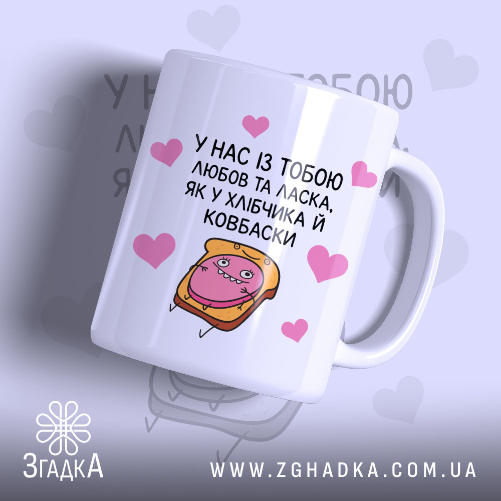 Chashka Khlibchyk I Kovbaska Idealnyi Podarunok Dlia Hurmaniv 1 Чашка хлібчик й ковбаска, біла з кольоровим принтом, показана з правого ракурсу, на фоні світло-фіолетового кольору з сердечками, з малюнком хлібчика і ковбаски, ручка класична, край рівний, глянцева поверхня.