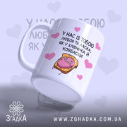 Чашка хлібчик й ковбаска, біла з кольоровим принтом, показана з лівого ракурсу, на фоні світло-фіолетового кольору з сердечками, з малюнком хлібчика і ковбаски, ручка класична, край рівний, глянцева поверхня.