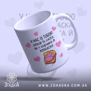 Чашка хлібчик й ковбаска, біла з кольоровим принтом, показана з легким нахилом, на фоні світло-фіолетового кольору з сердечками, з малюнком хлібчика і ковбаски, ручка класична, край рівний, глянцева поверхня.