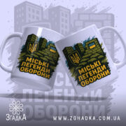 Чашка міські легенди оборони, дві чашки, біла, зображені з лівого ракурсу, на фоні стилізованих будівель, принт з гербом України та написом на чорному фоні, ручки класичні, краї рівні, глянцеві.