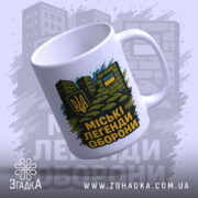 Чашка міські легенди оборони, біла, зображена з лівого ракурсу, на фоні стилізованих будівель, принт з гербом України та написом на чорному фоні, ручка класична, край рівний, глянцевий.
