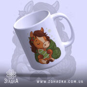 Чашка новорічний коник подарунок, біла, зображення коника з зеленим светром, тримає ялинку, ручка кругла, край рівний, глянцева поверхня, фон світло-синій.