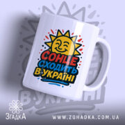 Чашка сонце в Україні, біла, з ракурсом зліва, на фоні світло-фіолетового кольору, з яскравим принтом сонця та написом 'СОНЦЕ СХОДИТЬ В УКРАЇНІ', ручка гладка, край рівний, глянцевий.