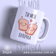 Чашка ти моя булочка, біла з малюнком двох усміхнених шматочків хліба, написом 'ТИ МОЯ БУЛОЧКА', фон світло-фіолетовий, ручка округла, край рівний, глянцева поверхня.