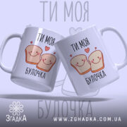 Чашка ти моя булочка, дві чашки, обидві білих з малюнком двох усміхнених шматочків хліба, написом 'ТИ МОЯ БУЛОЧКА', фон світло-фіолетовий, ручки округлі, краї рівні, глянцева поверхня.