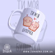 Чашка ти моя булочка, біла з малюнком двох усміхнених шматочків хліба, написом 'ТИ МОЯ БУЛОЧКА', фон світло-фіолетовий, ручка округла, край рівний, глянцева поверхня, нахилена вбік.