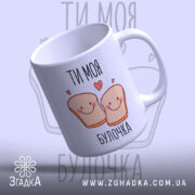 Чашка ти моя булочка, біла з малюнком двох усміхнених шматочків хліба, написом 'ТИ МОЯ БУЛОЧКА', фон світло-фіолетовий, ручка округла, край рівний, глянцева поверхня, нахилена вбік, видно внутрішню частину.