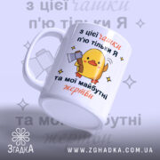 Чашка жорсткий жарт офісу: біла чашка з жовтим каченям, що тримає сокиру, на фоні світло-фіолетового кольору, текст на українській мові, ручка кругла, край гладкий, матова поверхня.