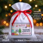 Дитячий святковий мішечок із написом “Від Святого Миколая” та червоною стрічкою на фоні ялинкових вогників.