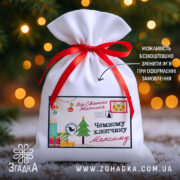 Дитячий святковий мішечок із написом “Від Святого Миколая” та червоною стрічкою на фоні ялинкових вогників.