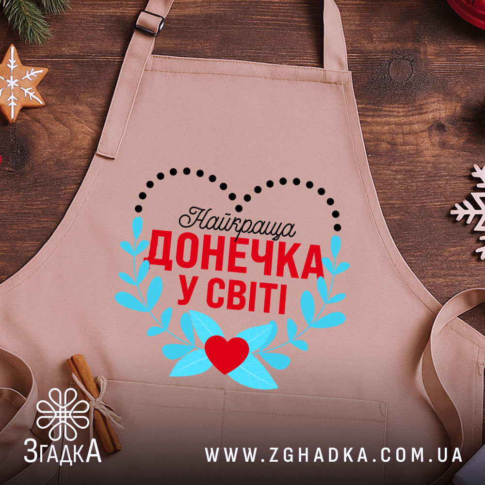 Фартух для донечки крупним планом на дерев’яній поверхні поруч із новорічним декором.