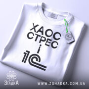 Футболка 1с хаос стрес, складена футболка на світлому фоні, ракурс зверху, принт чорний з текстом 'ХАОС СТРЕС i 1С', на комірці зелена етикетка.