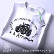 Футболка а я агроном. Біла футболка, складена на поверхні, з видимим принтом. Зображення з верхнього ракурсу. На фоні світло-сірий градієнт. Принт чорного кольору з трактором та написом 'ВСІ ЛЮДИ ЯК ЛЮДИ, А Я АГРОНОМ', з етикеткою зеленого кольору.
