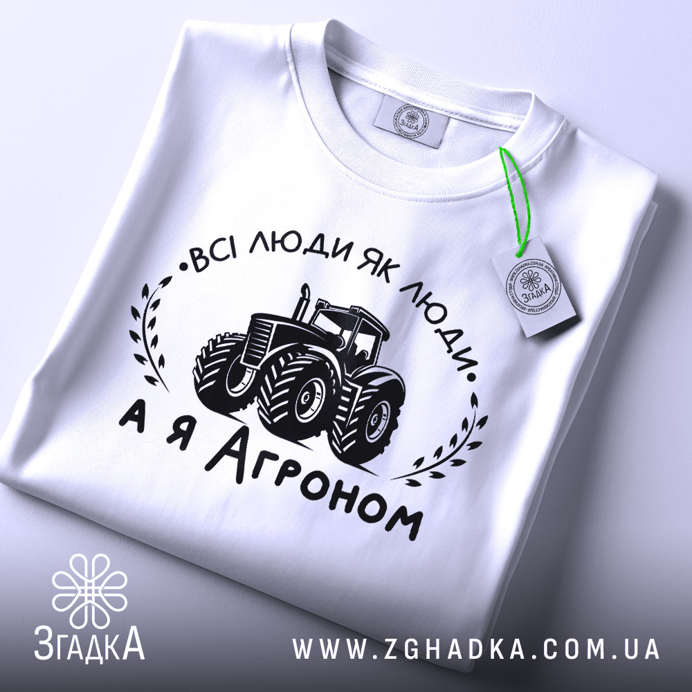 Футболка а я агроном. Біла футболка, складена на поверхні, з видимим принтом. Зображення з верхнього ракурсу. На фоні світло-сірий градієнт. Принт чорного кольору з трактором та написом 'ВСІ ЛЮДИ ЯК ЛЮДИ, А Я АГРОНОМ', з етикеткою зеленого кольору.