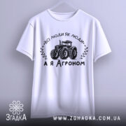 Футболка а я агроном. Біла футболка з коротким рукавом, підвішена на вішалці, зображена з фронтального ракурсу. На фоні світло-сірий градієнт. Принт чорного кольору з трактором та написом 'ВСІ ЛЮДИ ЯК ЛЮДИ, А Я АГРОНОМ'.