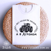 Футболка а я агроном. Біла футболка, складена на плетеній основі, зображена з верхнього ракурсу. На фоні світло-сірий градієнт. Принт чорного кольору з трактором та написом 'ВСІ ЛЮДИ ЯК ЛЮДИ, А Я АГРОНОМ'.