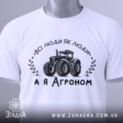 Футболка а я агроном. Біла футболка, складена на поверхні, з видимим принтом. Зображення з верхнього ракурсу. На фоні світло-сірий градієнт. Принт чорного кольору з трактором та написом 'ВСІ ЛЮДИ ЯК ЛЮДИ, А Я АГРОНОМ', з етикеткою зеленого кольору.