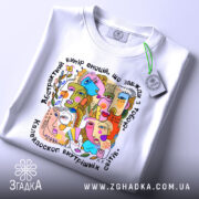 Футболка абстрактні емоції, біла, зображена зверху на столі, фон світло-сірий, принт кольоровий, з елементами абстрактних облич, на футболці є етикетка з зеленим шнурком.