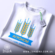 Футболка агроном син землі, біла, складена на поверхні, зображена з верхнього ракурсу, фон світлий, принт з жовтими колосками та синьою стрічкою з написом "АГРОНОМ", на ярлику зелена стрічка.